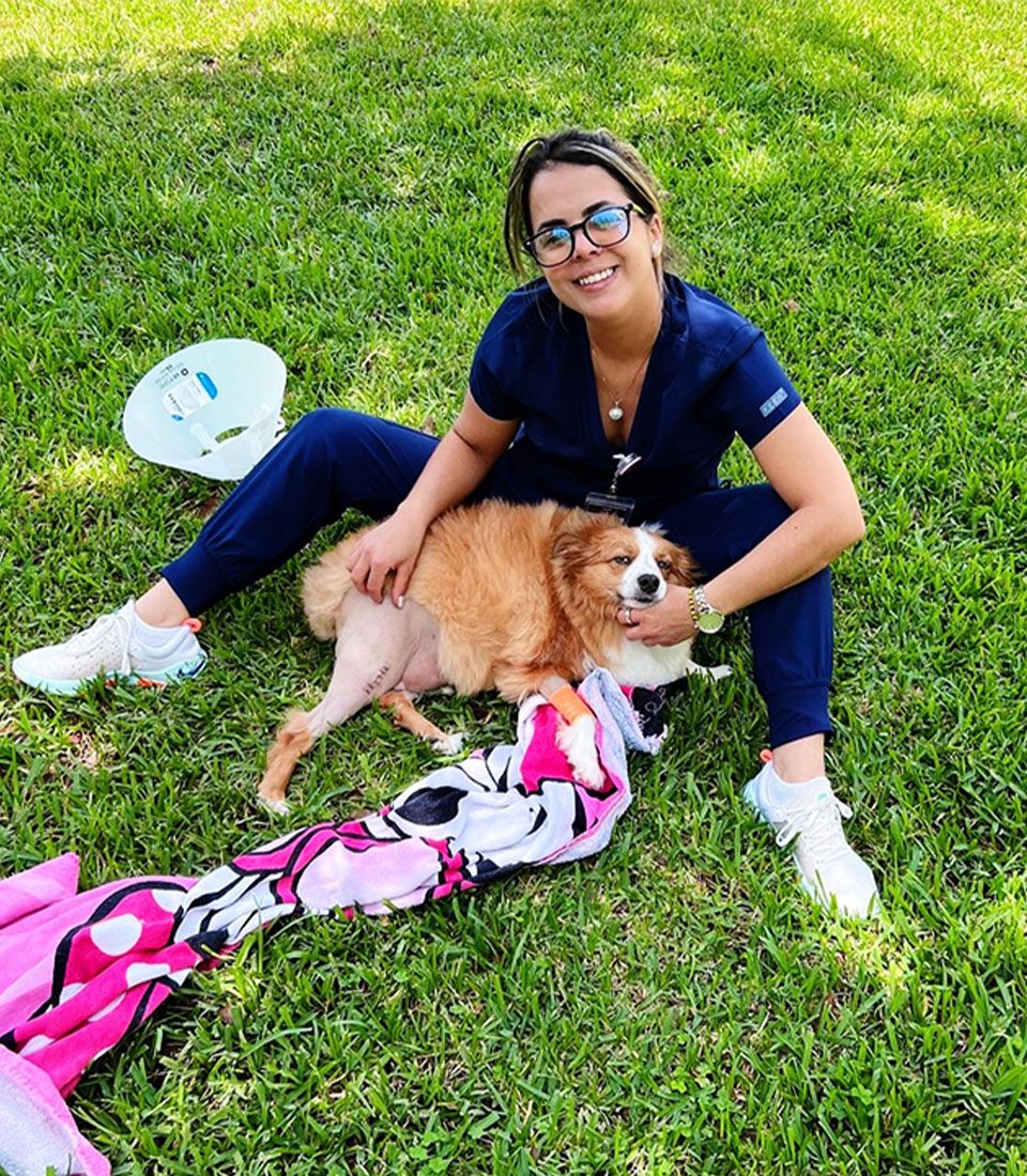 Person in blue scrubs sitting on grass, smiling, with a fluffy brown and white dog lying on their lap.