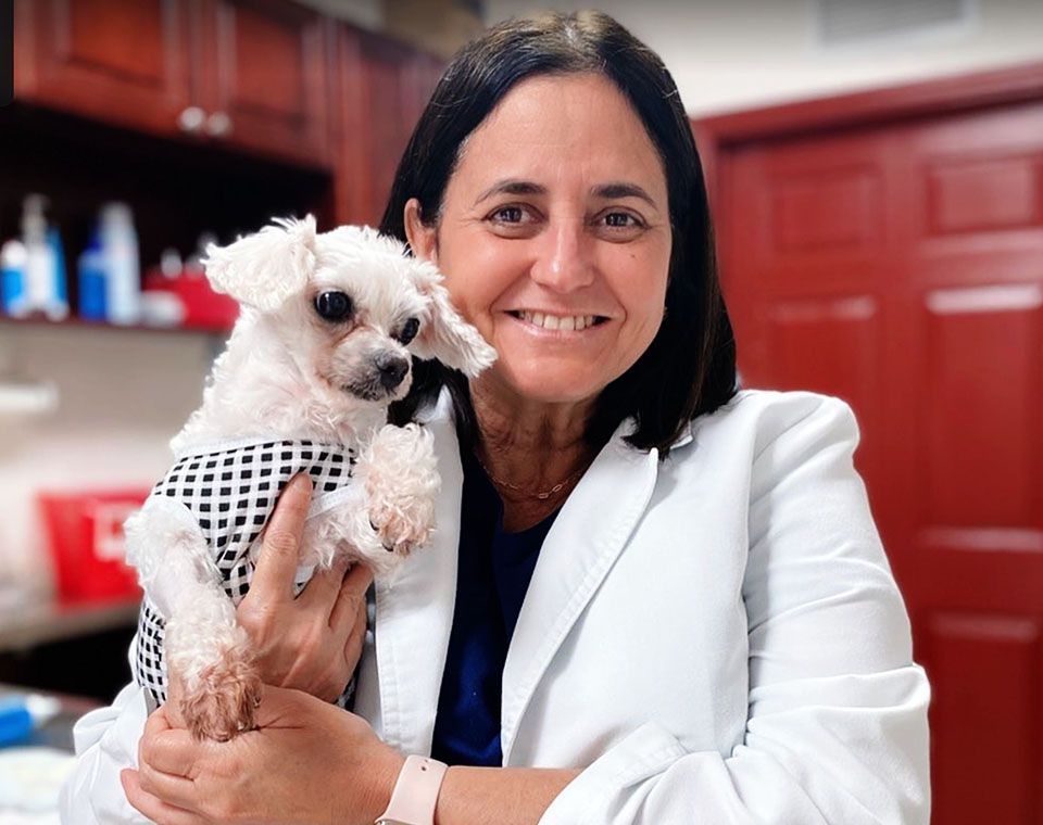 A woman in a white coat smiles while holding a small white dog wearing a checkered outfit.