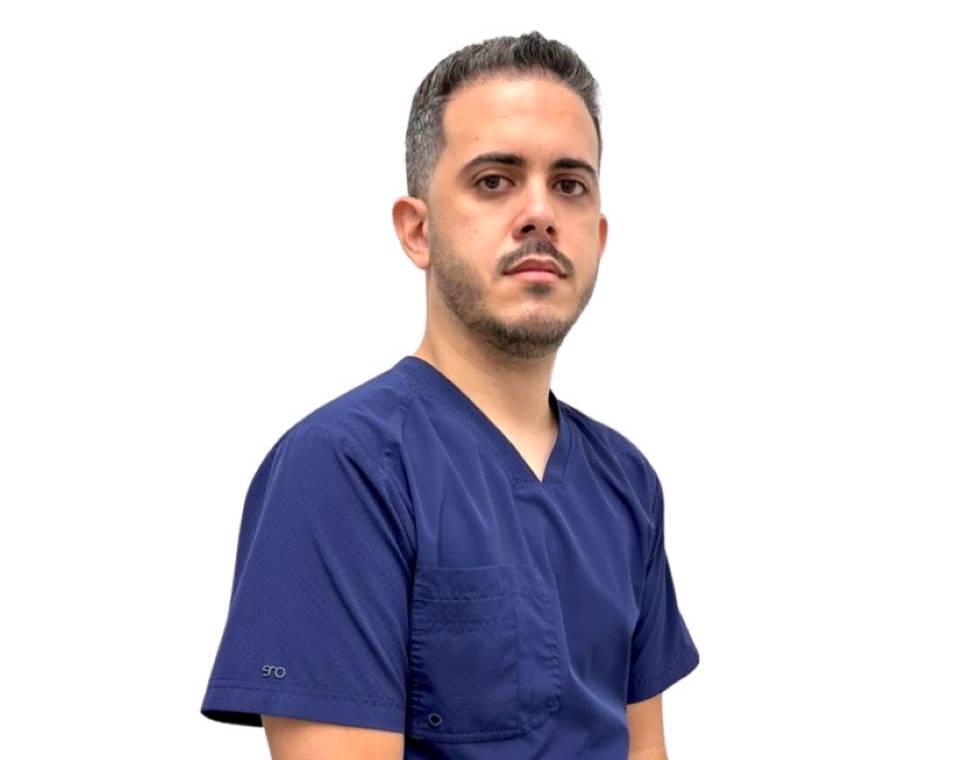A man with short hair and a beard wearing a blue scrub top, looking forward.
